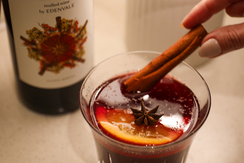 Close-up of woman’s hand stirring a spice mocktail with a stick of cinnamon. A bottle of Edenvale mulled wine in the background.