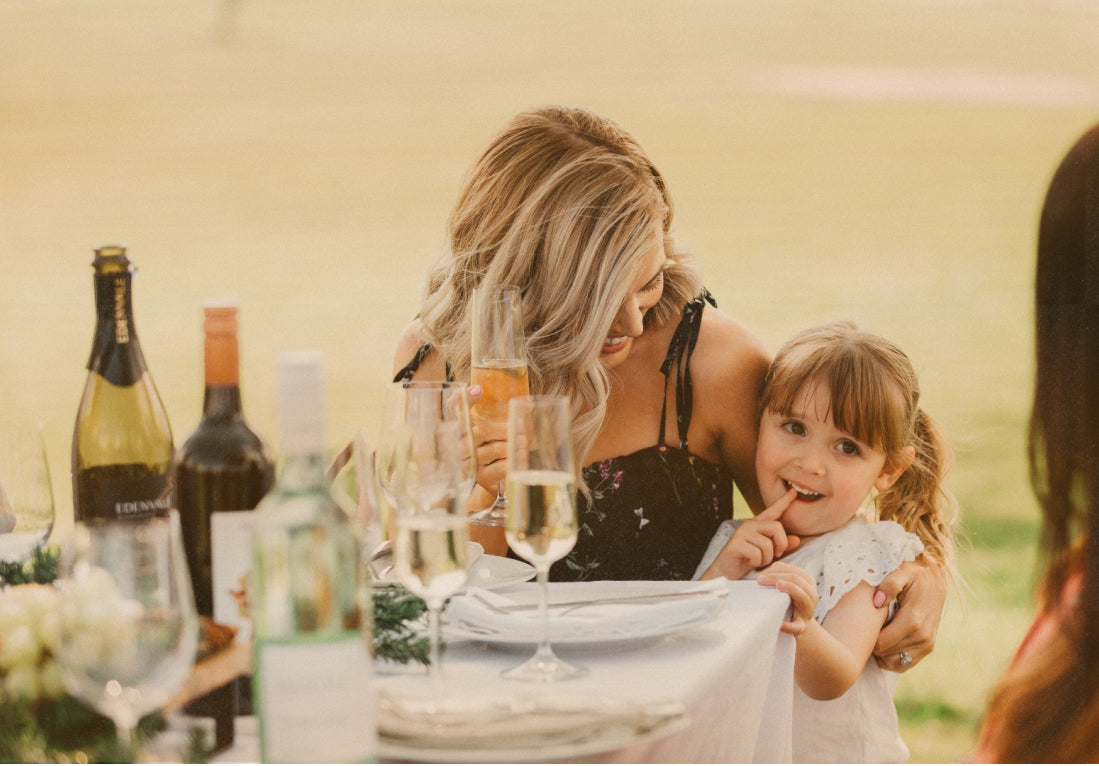 Woman sitting at table holding a glass of non-alcoholic rose in one hand and cuddling young girl standing by her side