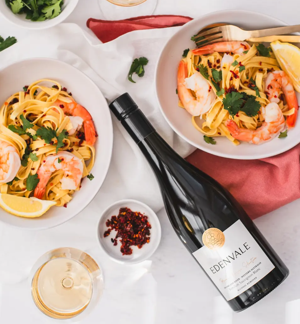 Aerial shot of bottle of Edenvale non-alcoholic white lying on a white tablecloth alongside prawn pasta dishes and bowl of chilli flakes