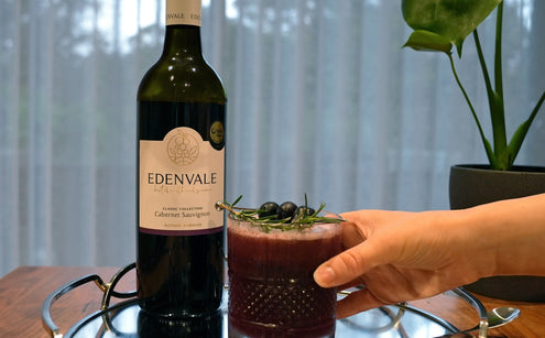  Black Cedar Sour Mocktail in a short glass garnished with blackcurrants and rosemary beside a bottle of Edenvale Cabernet Sauvignon. 