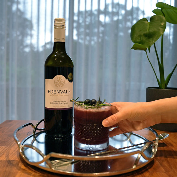  Black Cedar Sour Mocktail in a short glass garnished with blackcurrants and rosemary beside a bottle of Edenvale Cabernet Sauvignon. 