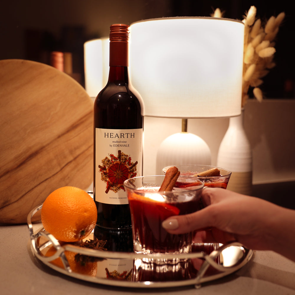 Woman’s hand around a Heart & Stone mocktail on a silver tray, with bottle of Edenvale Hearth and mood lighting in background