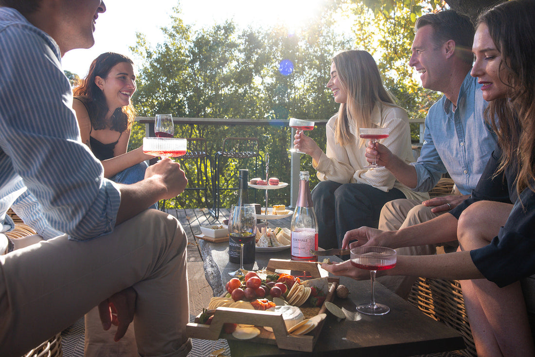 Group of friends gathered around outdoor table drinking non-alcoholic wine and laughing 