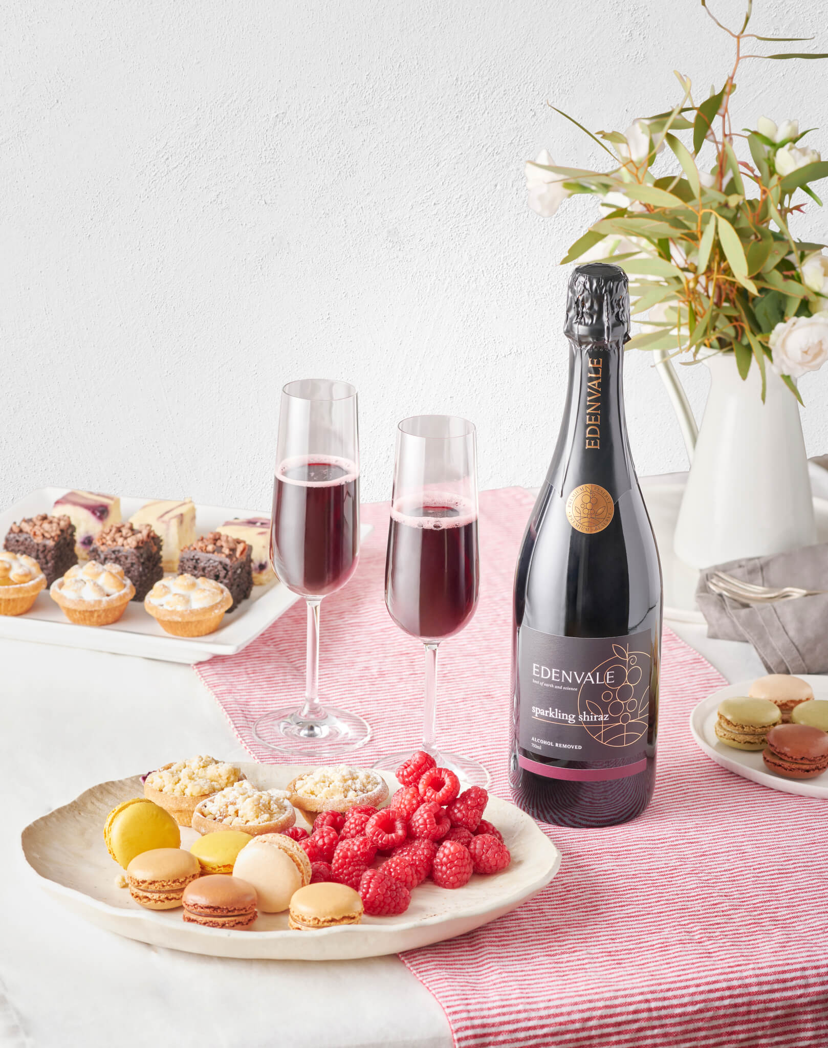 A bottle and two Champagne flutes full of Non-alcoholic Sparkling Shiraz on a pink table runner with plates of colourful macarons, mini sweet tarts and fresh raspberries.