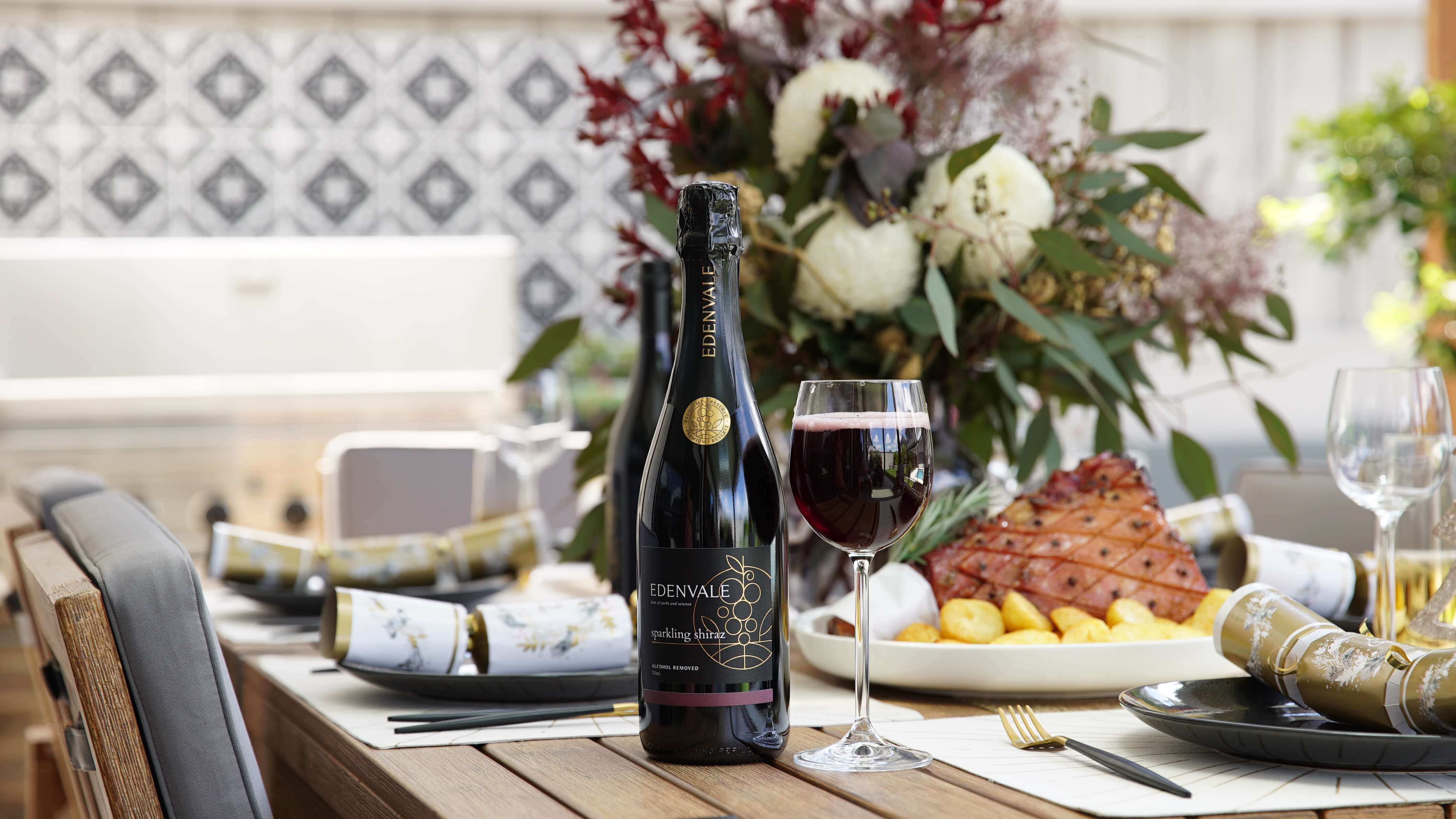Bottle of Edenvale Sparkling Shiraz on a table with Christmas crackers, a shared platter of glazed ham with roast potatoes, and a vase of Australian flora.