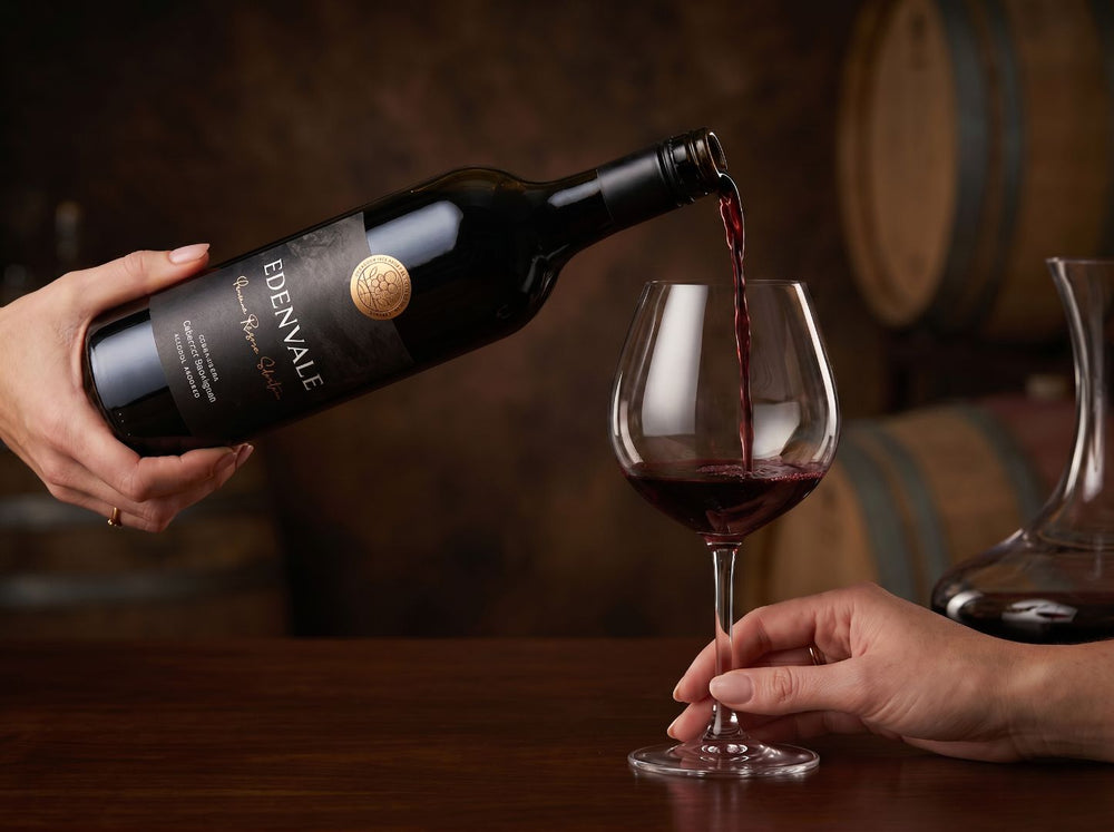 Close-up of a hand pouring from a bottle of Edenvale Premium Cabernet Sauvignon into a large red wine glass. Dark, moody background.