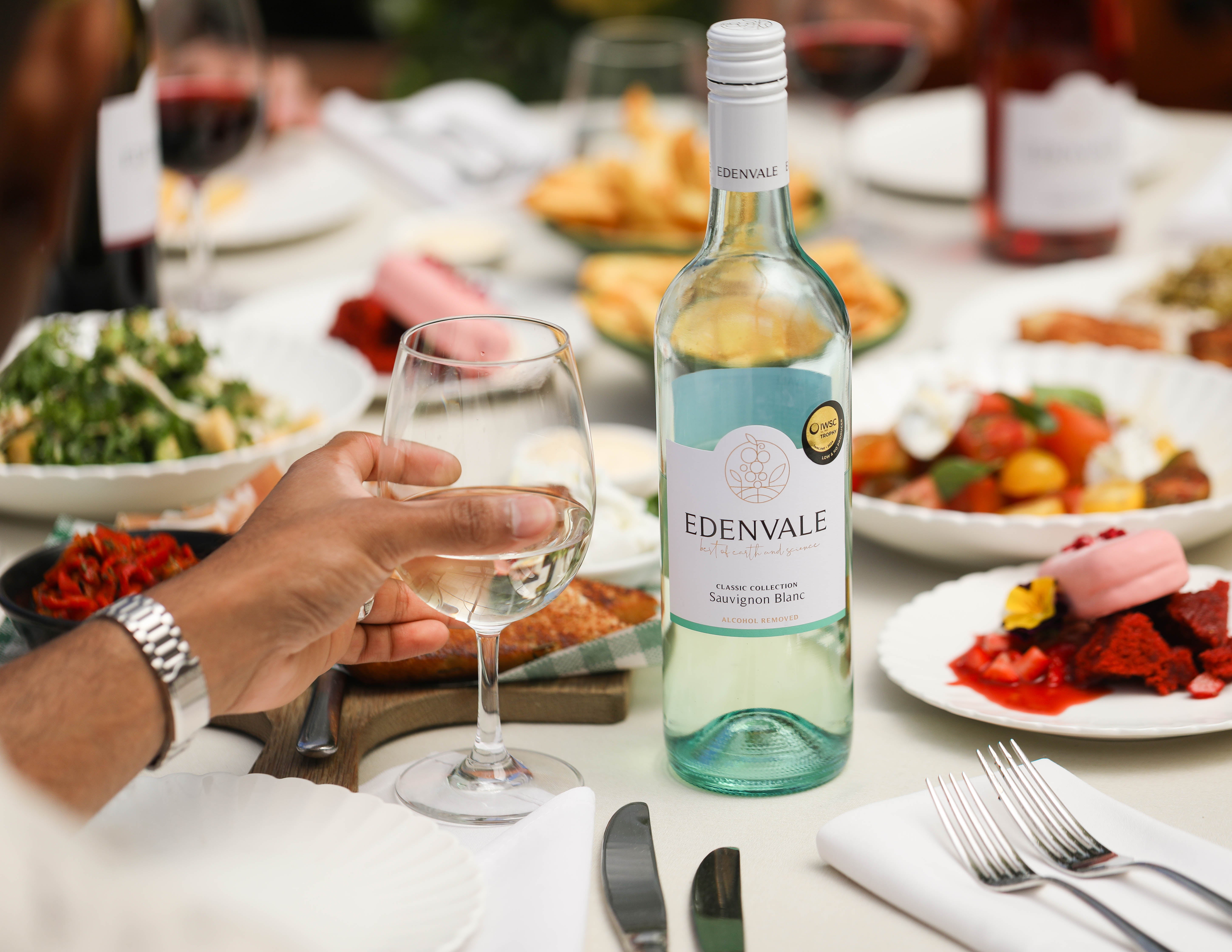 Edenvale Non-Alcoholic Sauvignon Blanc bottle on a dining table set for a shared meal, with a hand holding a glass of white wine

