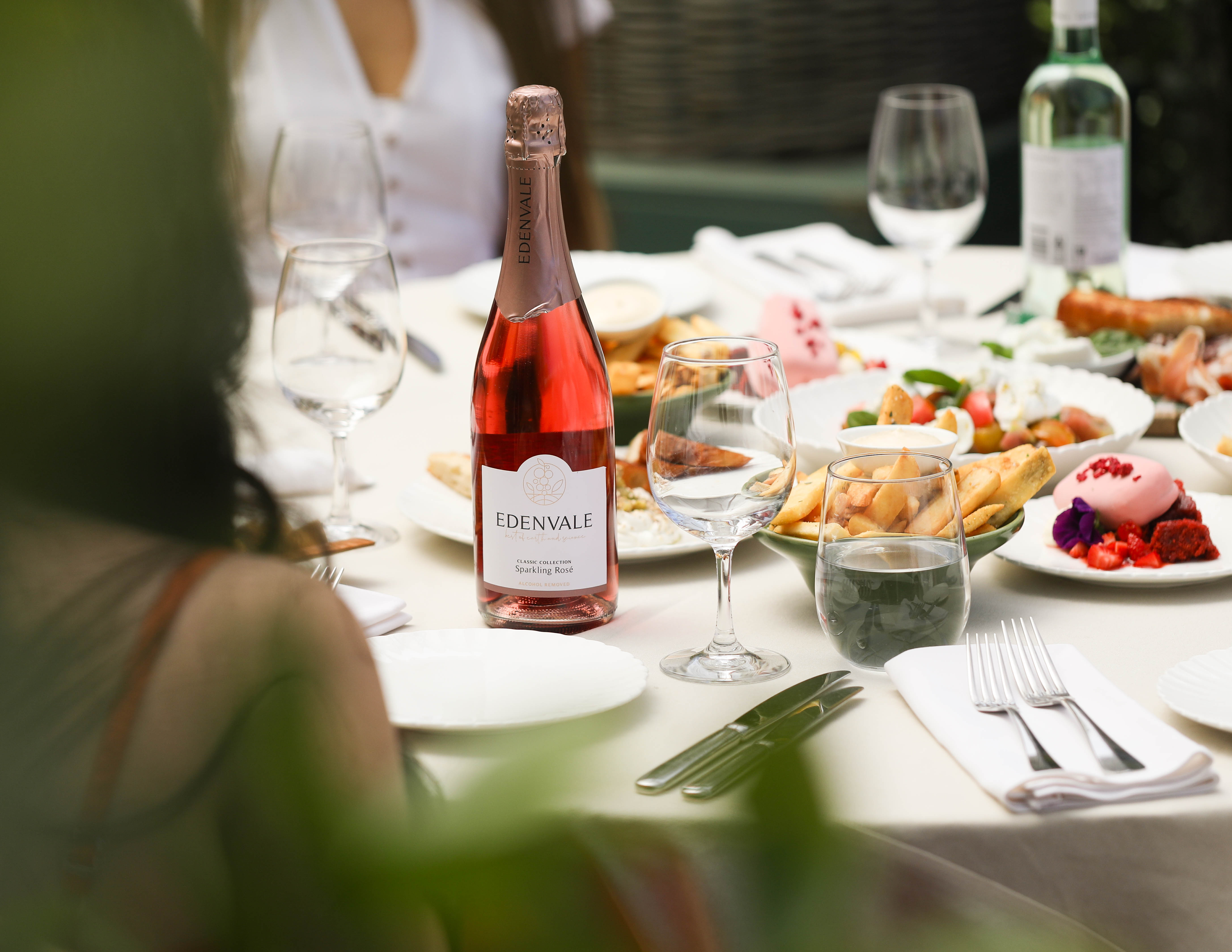 Edenvale Non-Alcoholic Sparkling Rosé bottle on an outdoor dining table alongside glasses, cutlery, and plates of fresh food.