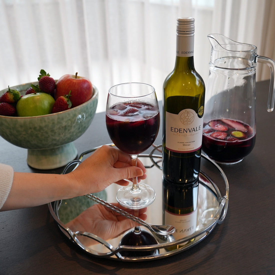 Hand holding a glass of Red Sangria made with Edenvale non-alcoholic wine, styled with fresh fruit and a serving carafe 
