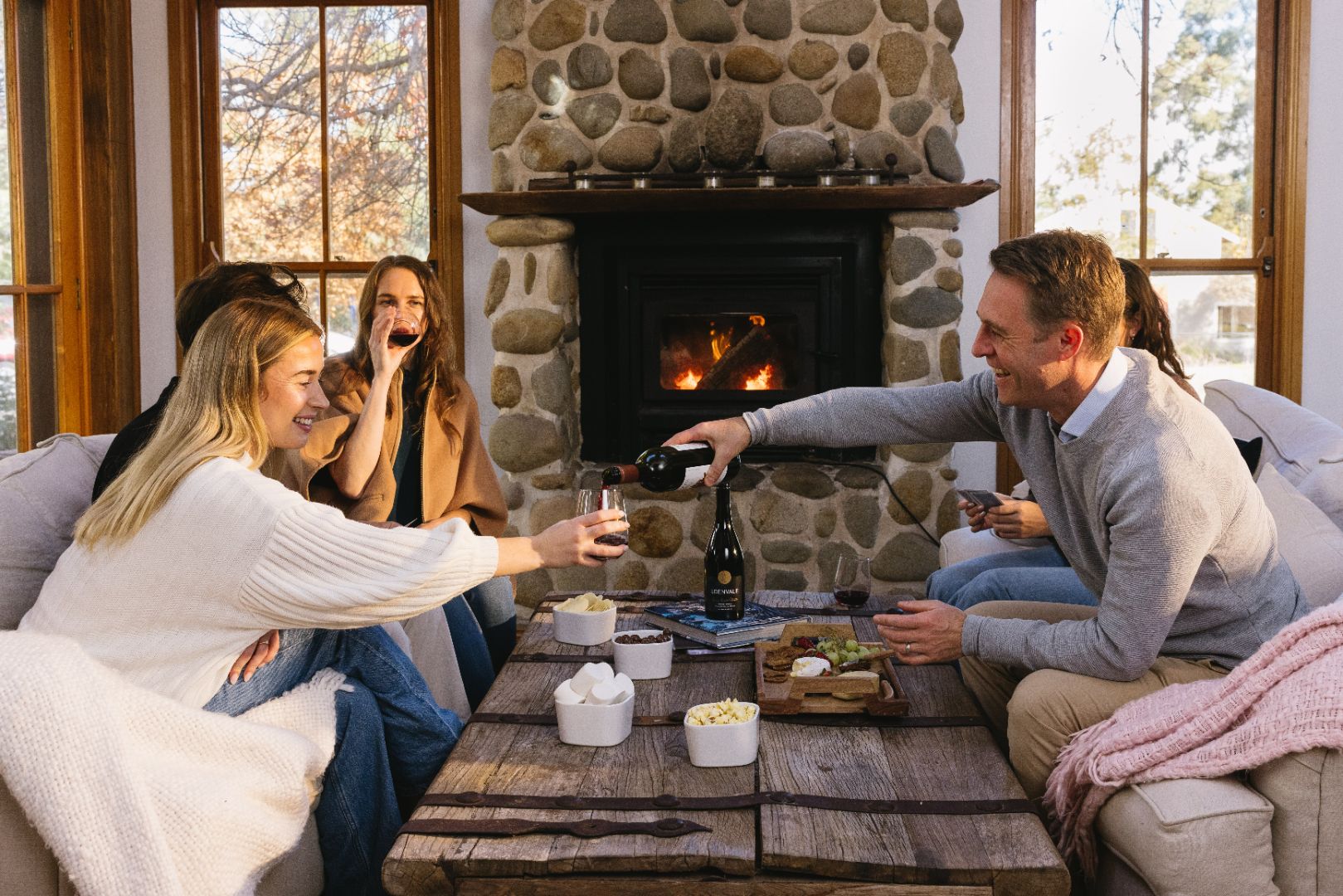 Friends gathered by a fireplace as a man pours Edenvale non-alcoholic red wine, with a grazing board on the table