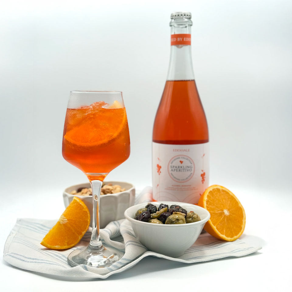 Full glass of bittersweet Sparkling Aperitivo with a wedge of orange. Appetisers, a cut orange and a full bottle are in the background